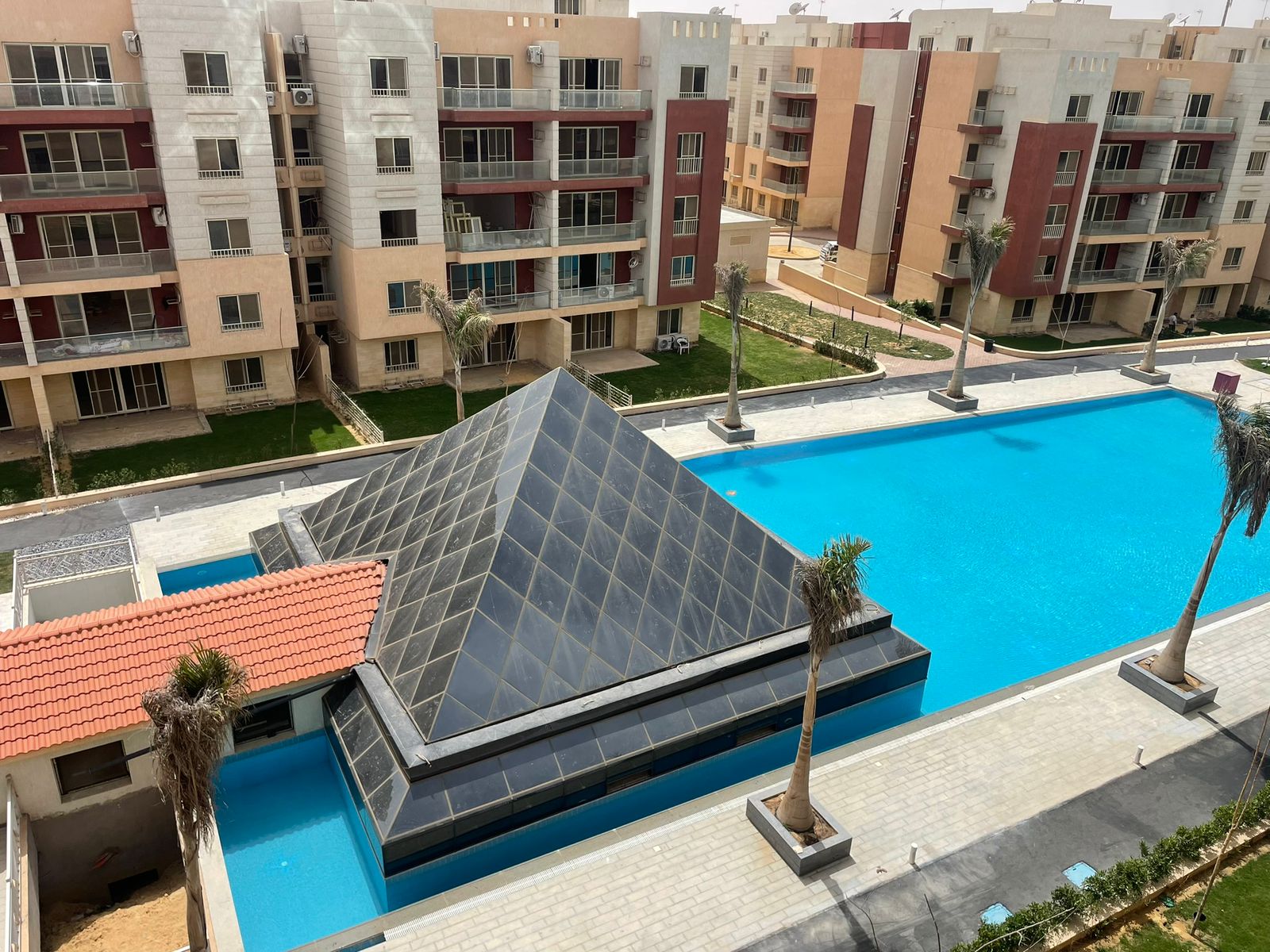 Pool with a glass pyramid roof and palm trees among apartment buildings - penthouse in Promenade, New Cairo