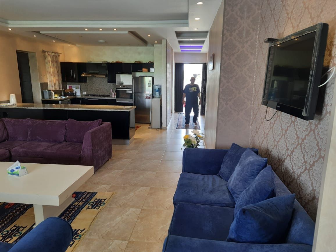 property-image-69f07468302ef مساحة معيشة مفتوحة بكنبة زرقاء وتلفزيون حائط بجوار المطبخ - شاليه في هاسيندا باي، سيدي عبدالرحمن الساحل الشمالي