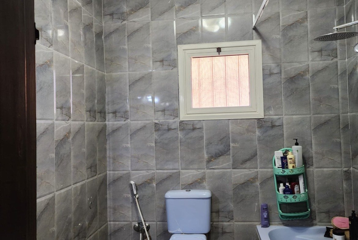 Bathroom with grey wall tiles, a corner bathtub and a small window - apartment in Al Narges, New Cairo