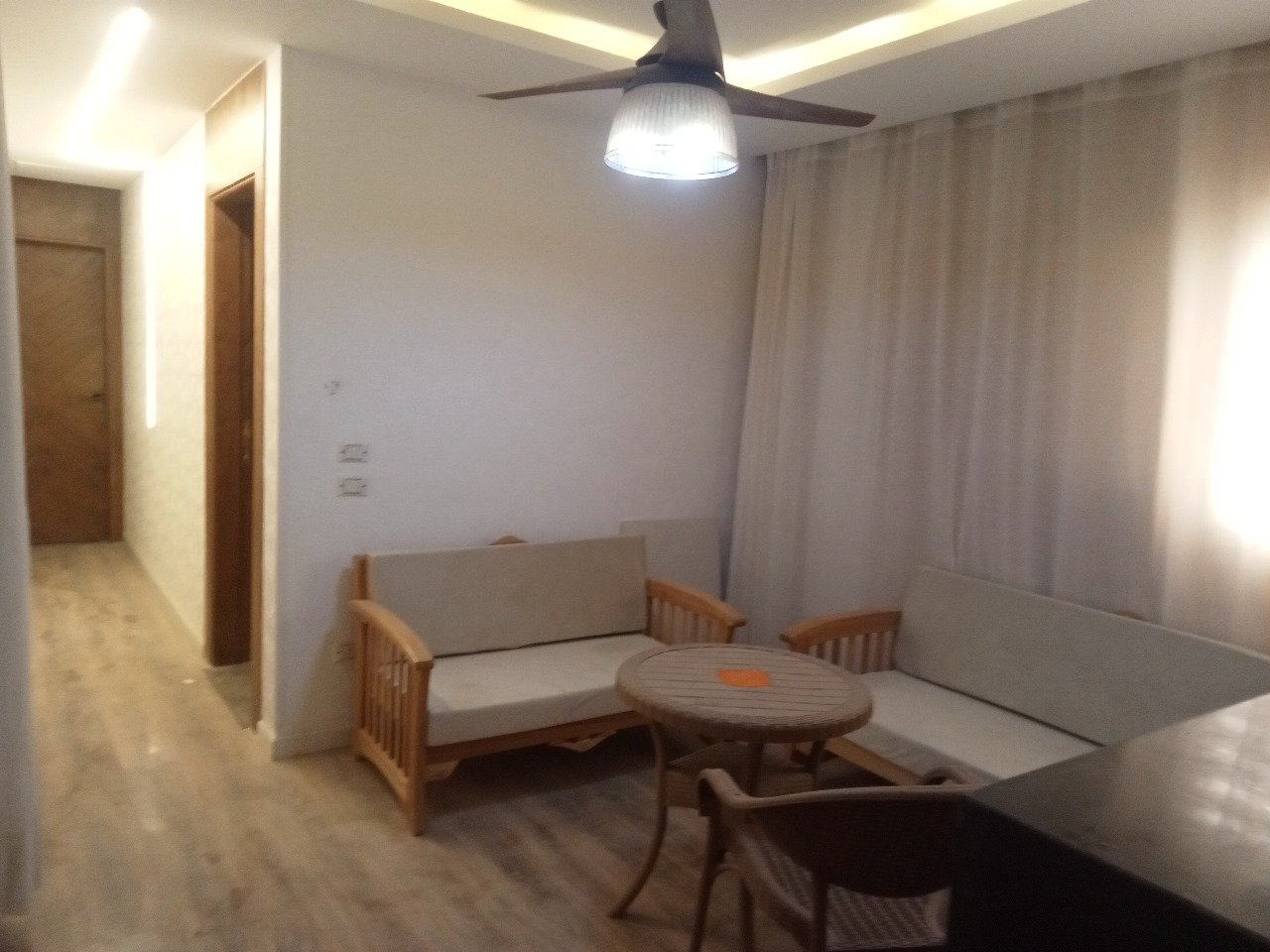 Compact living room with a ceiling fan, a wooden sofa and a round table - apartment in Sarai, Mostaqbal City