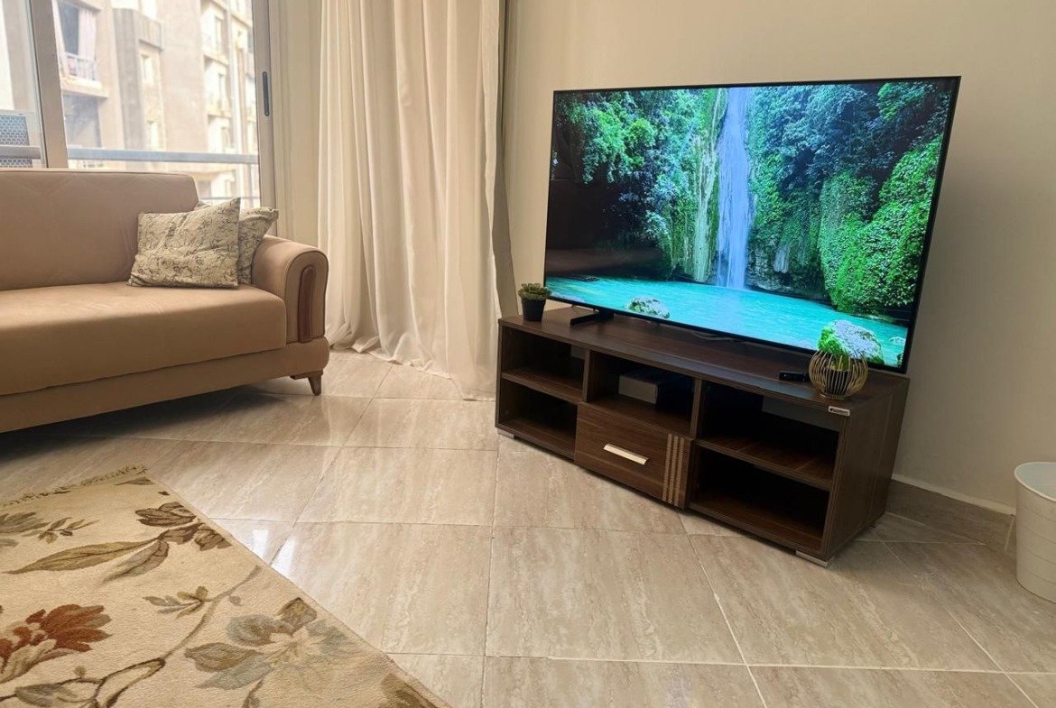 TV unit with a floral rug and a tan sofa corner - apartment in Dar Misr Al Koronfol New Cairo – 3 Bedrooms 130 sqm, New Cairo