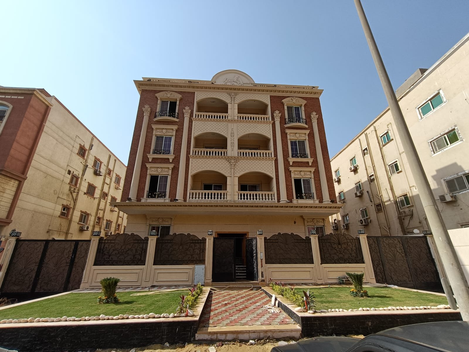 Wide front view of a terracotta apartment building with small lawns - apartment in Gharb Arabella, 5th Settlement