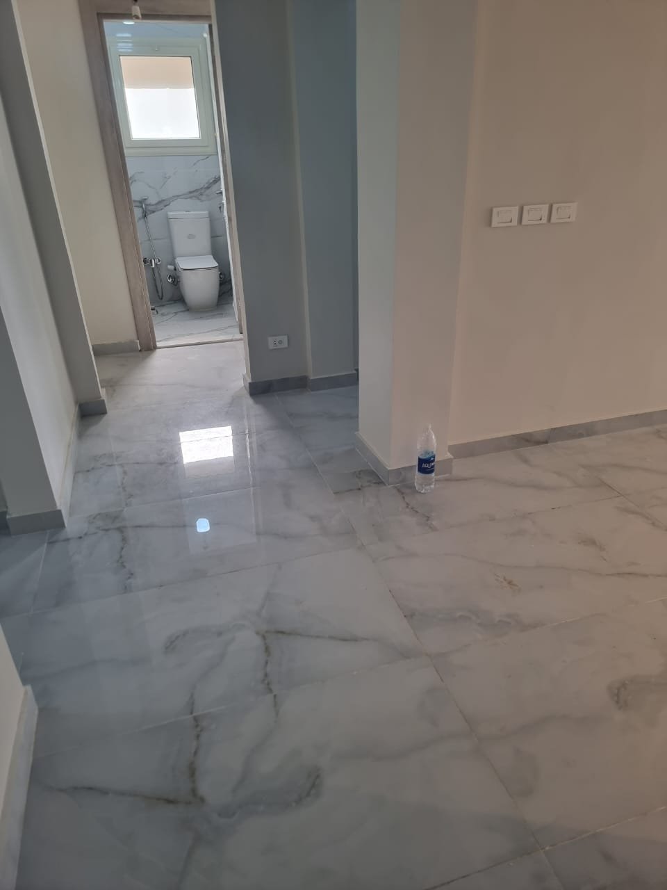 Hallway with white marble floors and a bathroom door in the distance - apartment in Green Square, Mostaqbal City