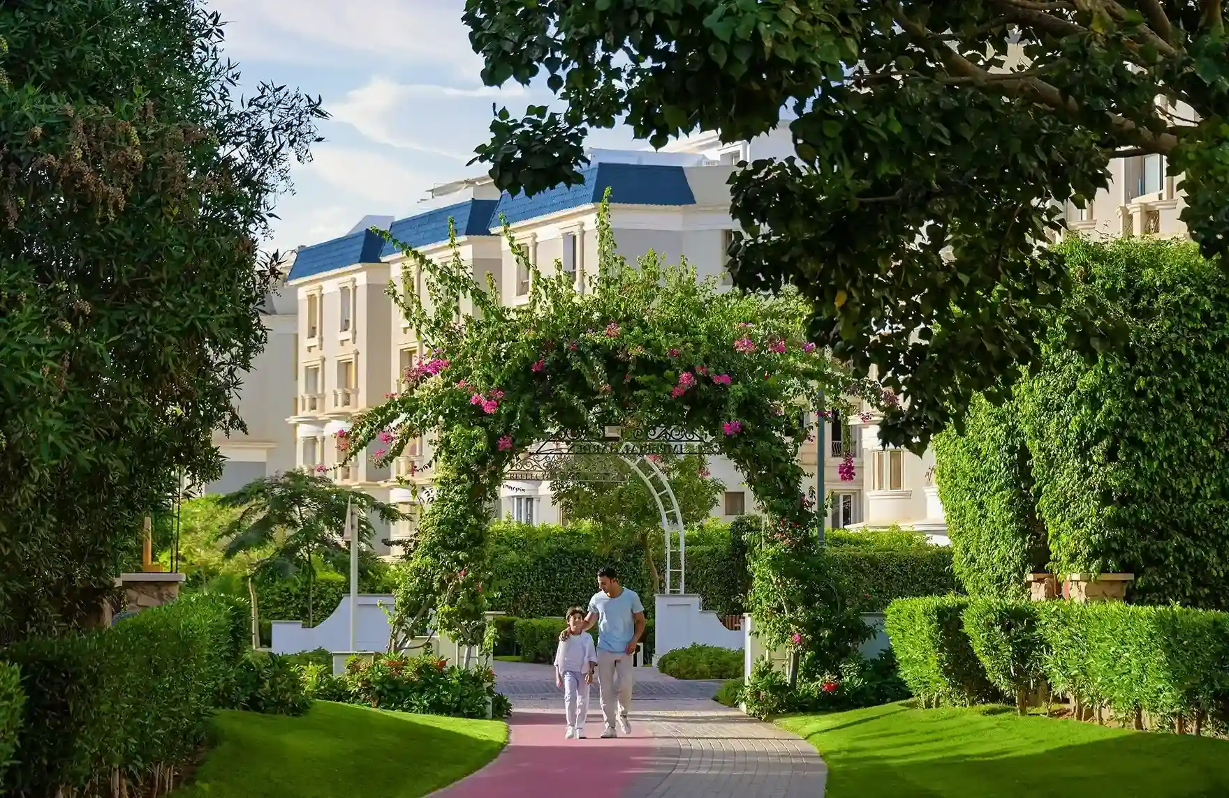 mountain-view-hyde-park-landscaped-walkways-general قوس حديقة مغطى بالزهور وممشى وردي يؤدي إلى مبانٍ بحواف زرقاء - فيلا في ماونتن فيو هايد بارك، التجمع الخامس